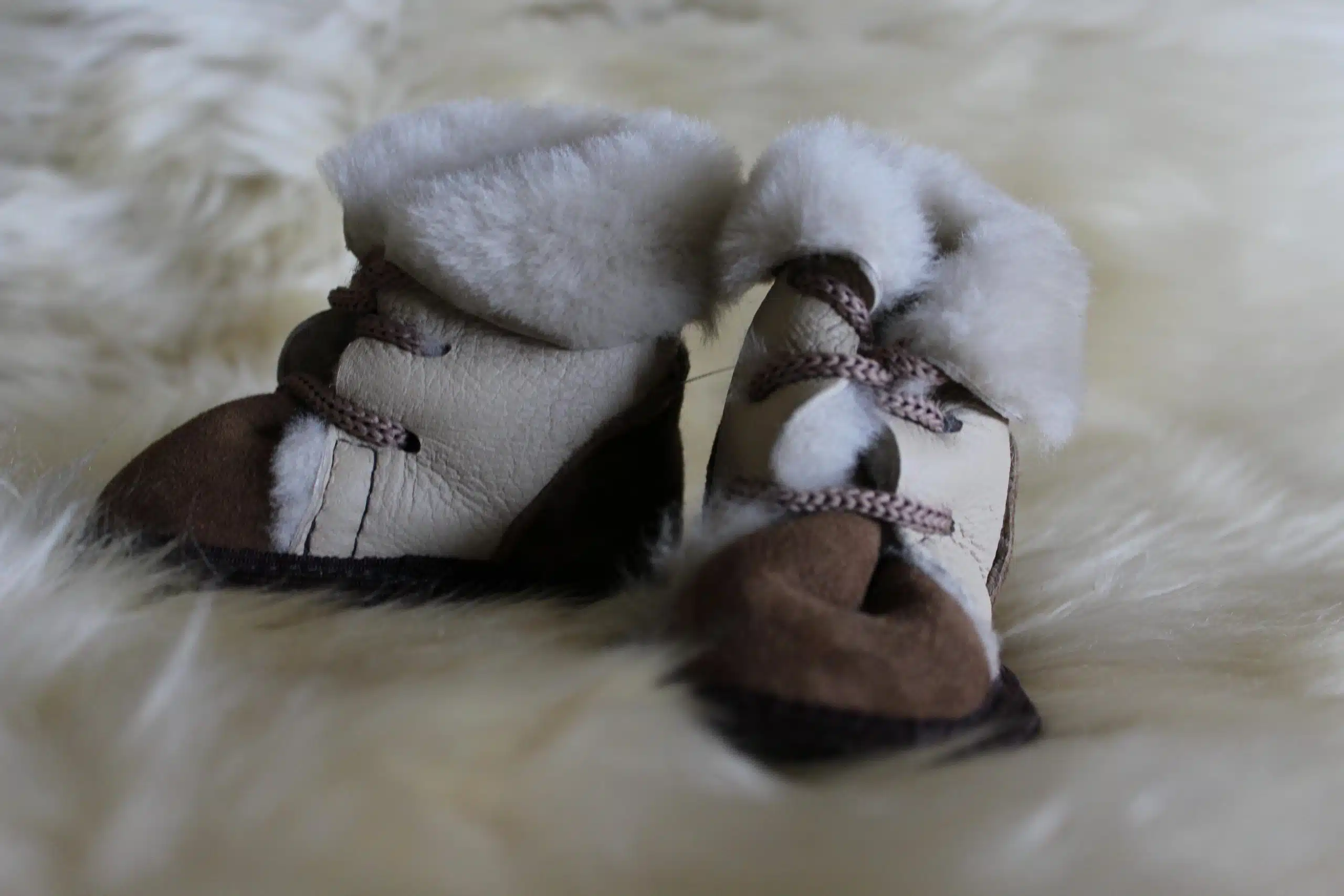 Ein Paar beige-braune Babyschuhe aus Leder mit weißem Fellbesatz, liegend auf flauschigem weißem Fell.